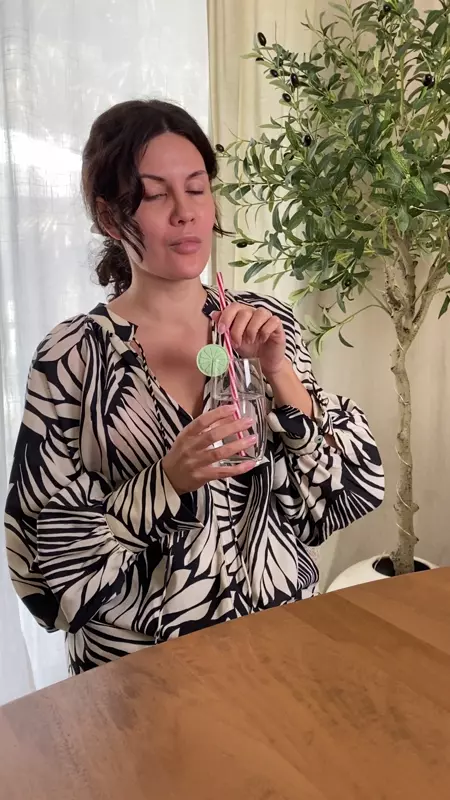 Woman with eyes closed holding glass with lemon slice and striped straw, next to olive tree indoors.