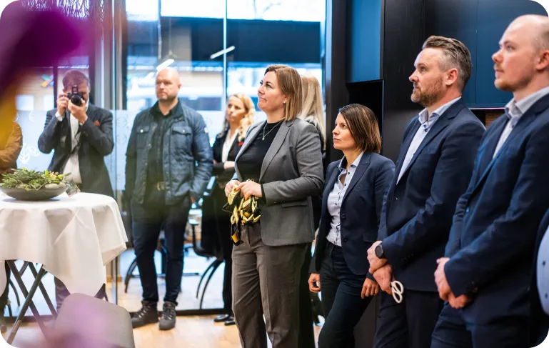 En gruppe voksne mennesker i formelle klær lytter oppmerksomt under et møte eller arrangement i et moderne kontormiljø.