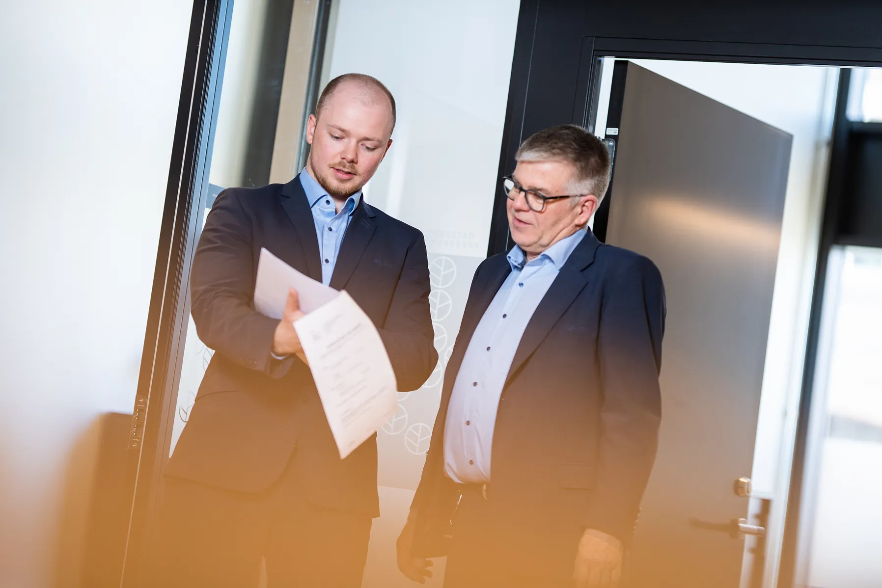 Two men in suits standing in an office doorway reviewing a document together.