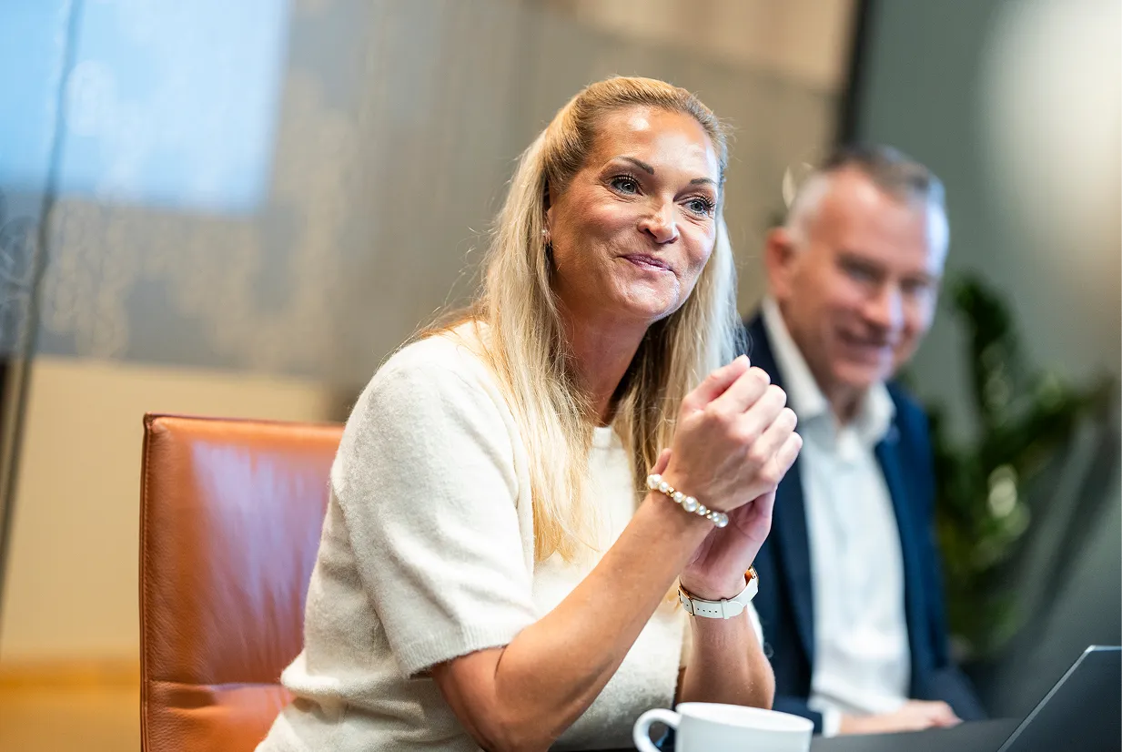 Kvinne med blondt hår som sitter ved et bord og snakker, med en mann i bakgrunnen som smiler.