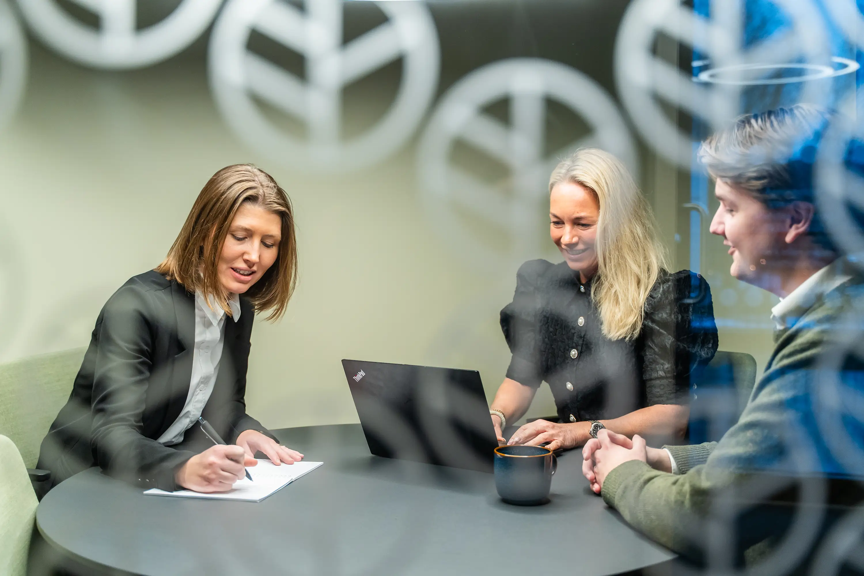 Tre personer i et møterom, en kvinne skriver notater, en annen bruker laptop, og en mann smiler.