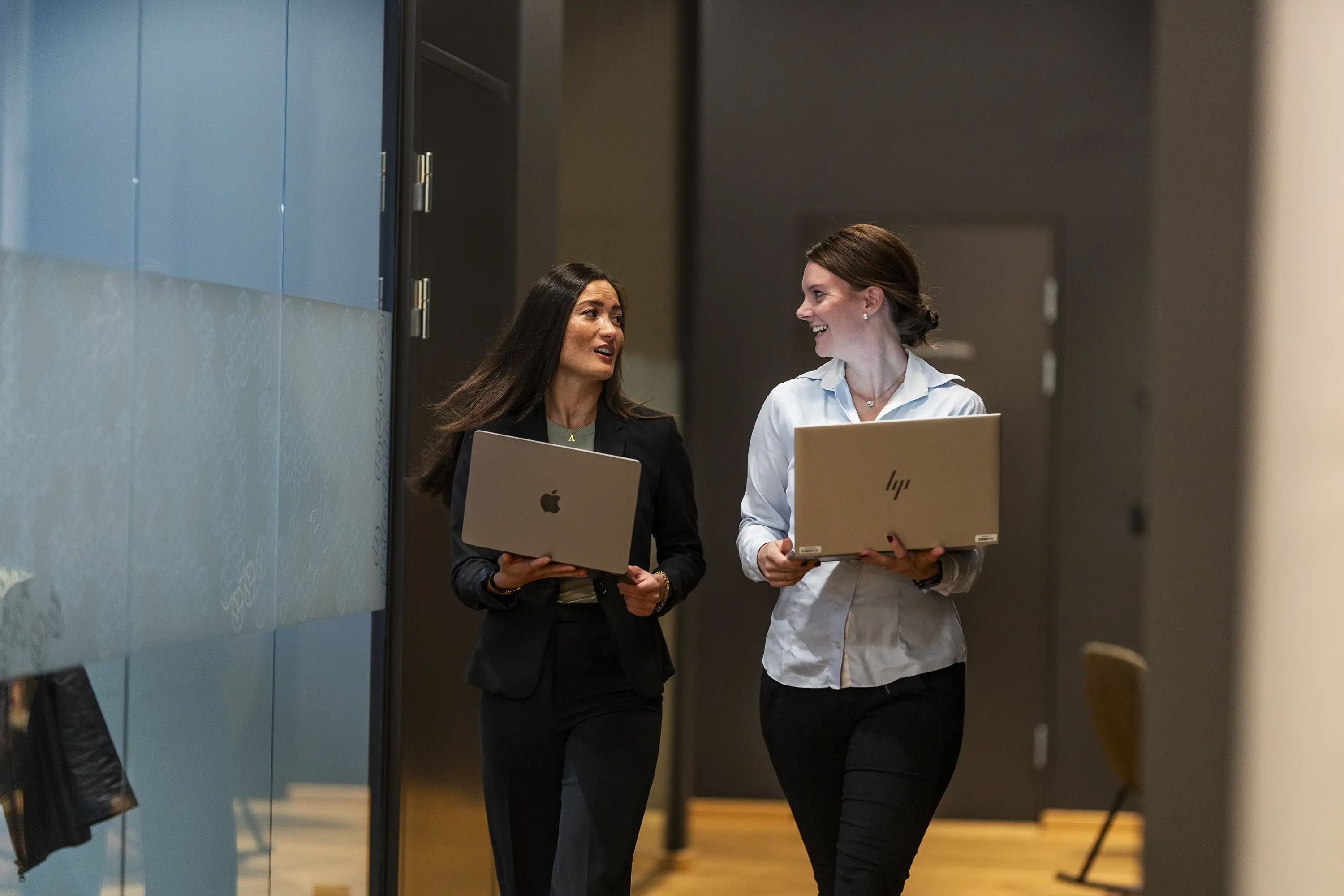 To profesjonelle kvinner går sammen i en kontorgang med bærbare datamaskiner og snakker.