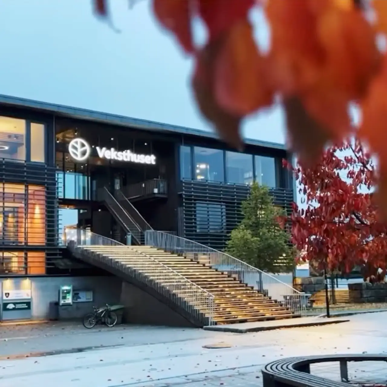 Moderne bygning med opplyst trapp og skiltet Veksthuset, omgitt av høstfargede trær.