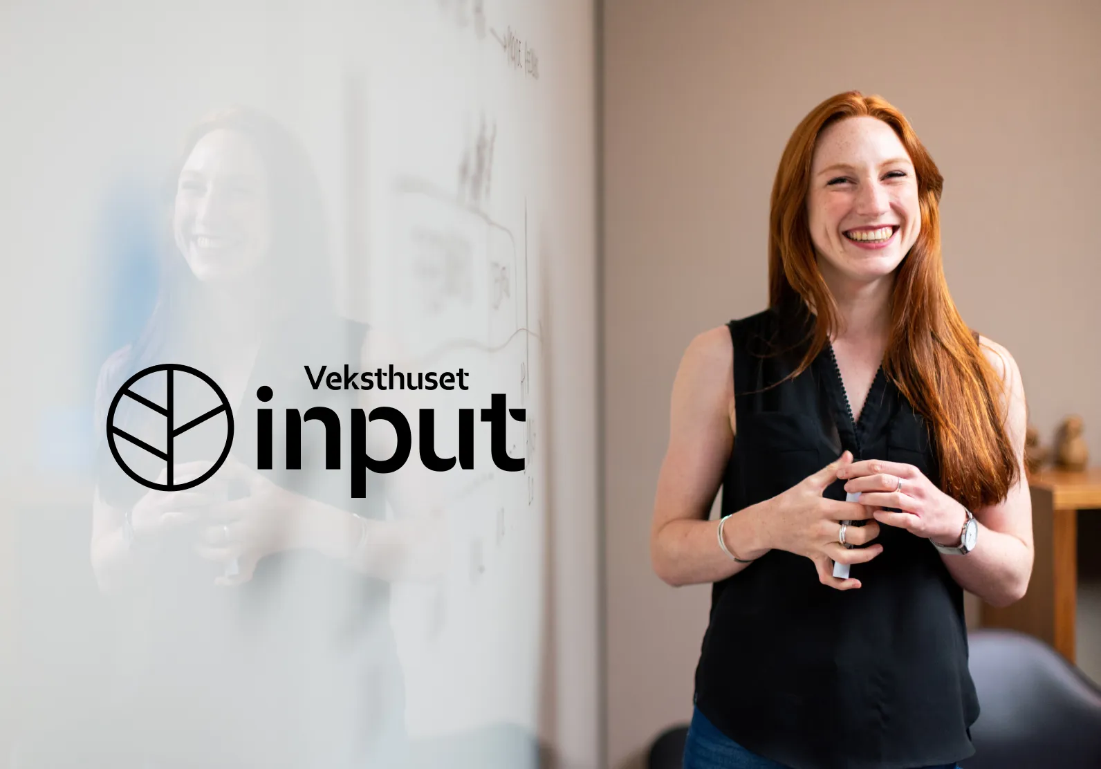 Smilende kvinne med rødt hår og svart ermeløs skjorte står ved en tavle med notater og holder en penn.