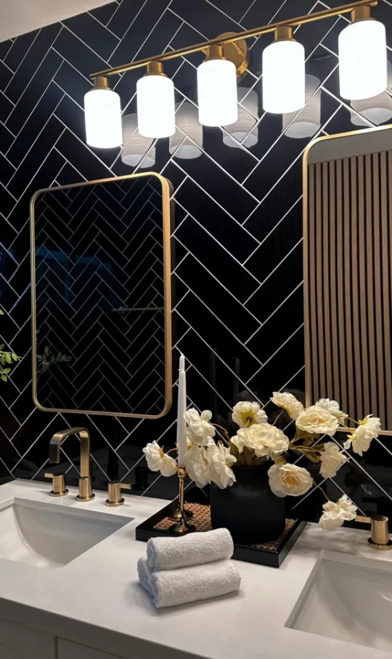 Black herringbone tile vanity wall with brass lighting and gold mirror