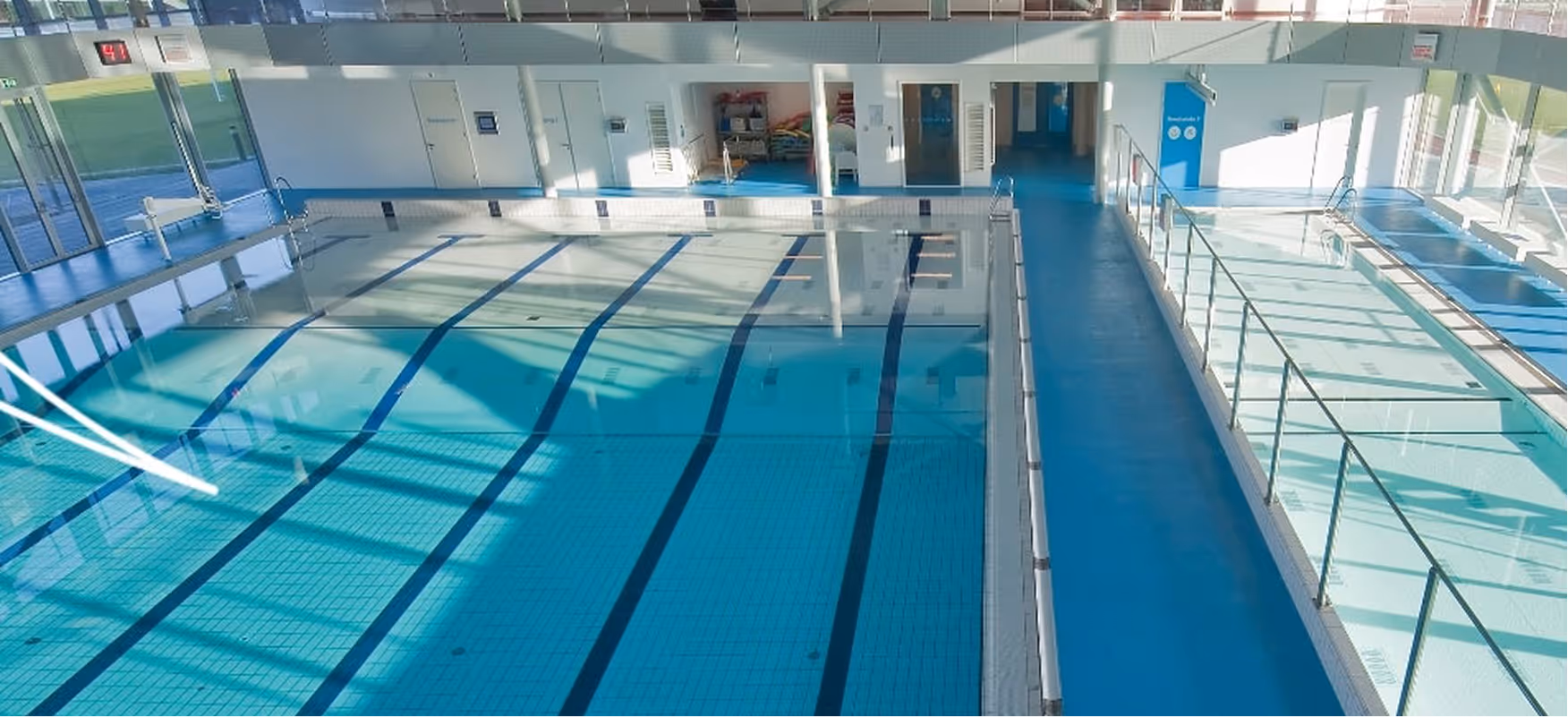 Indoor swimming pool area with empty lanes and adjacent shallow pools with metal railings.