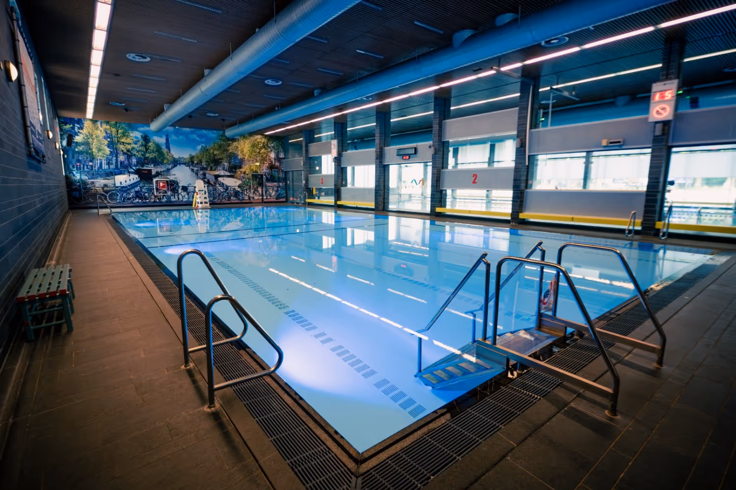 Indoor swimming pool with clear water, metal handrails, and a mural of a canal with bicycles on the far wall.