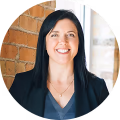 marketing for realtor - Smiling woman with straight black hair wearing a black blazer and gray top, standing in front of a brick wall.