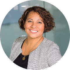 marketing for realtor - Smiling woman with curly short hair wearing a striped blazer and black top in front of a glass background.