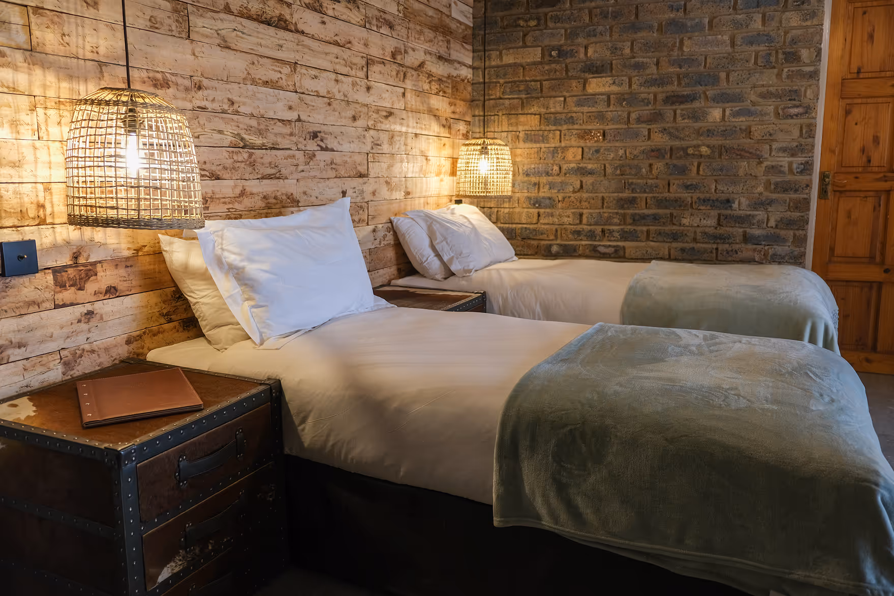 Cozy bedroom with two single beds, white bedding, green blankets, wooden and brick walls, and pendant rattan lamps.