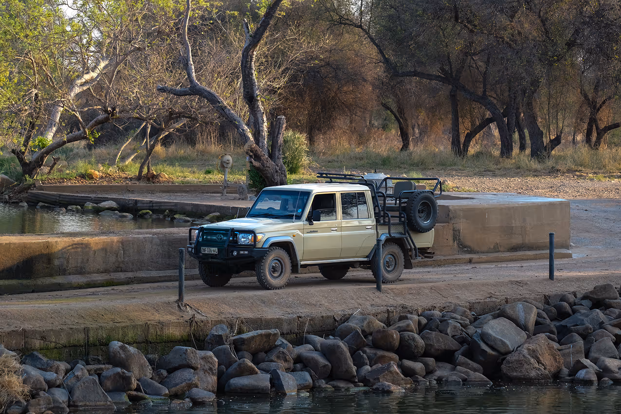 Beige safari vehicle parked on a dirt road near a water body with rocky shore and trees in the background.