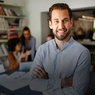 Lächelnder Mann mit verschränkten Armen im Büro, im Hintergrund Kollegen im Gespräch.