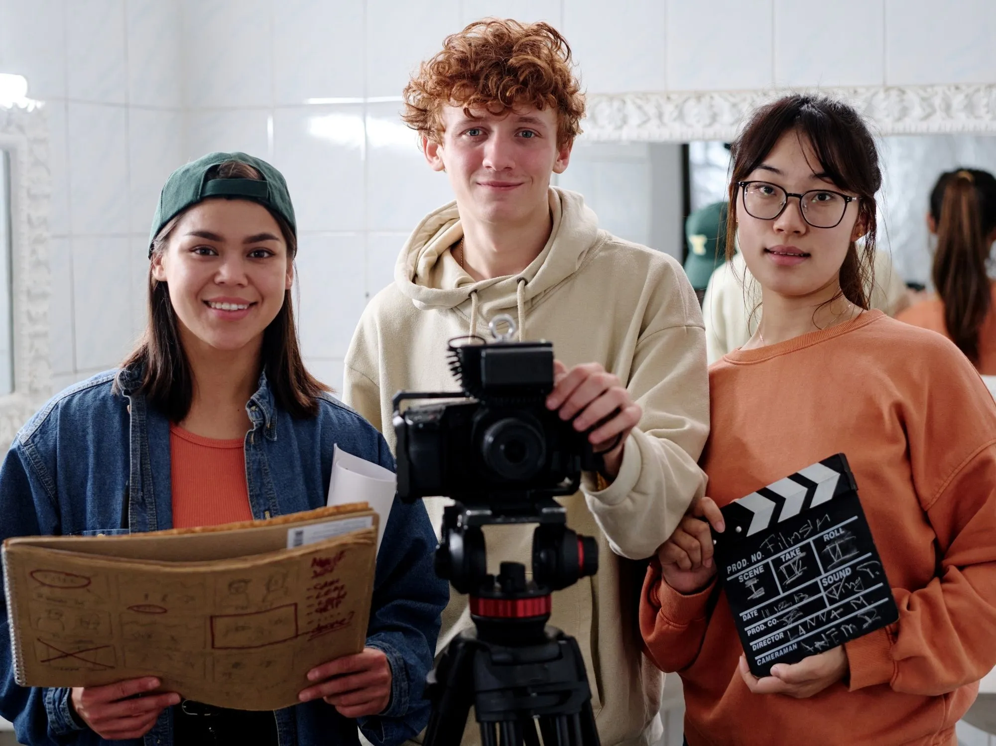 Three people in office: woman with newspaper, man with video camera, woman with clapperboard