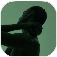 A person posing against a green background, turning their head and resting a hand on their shoulder.