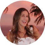 A person smiling broadly, standing outdoors during sunset near palm trees.