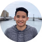 Young person smiling outdoors with water and buildings in the background.