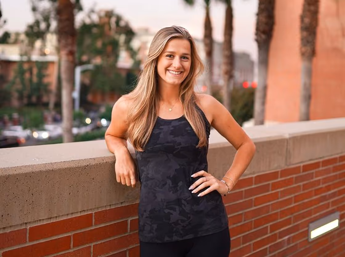 A person smiling and standing outdoors near a brick wall during the day.