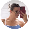 A person takes a mirror selfie, holding a red phone and wearing glasses.
