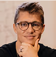 A person with glasses smiling, resting their chin on their hand.