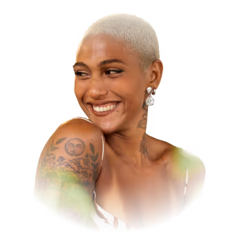A person with short hair and tattoos smiles, wearing earrings and a sleeveless top.