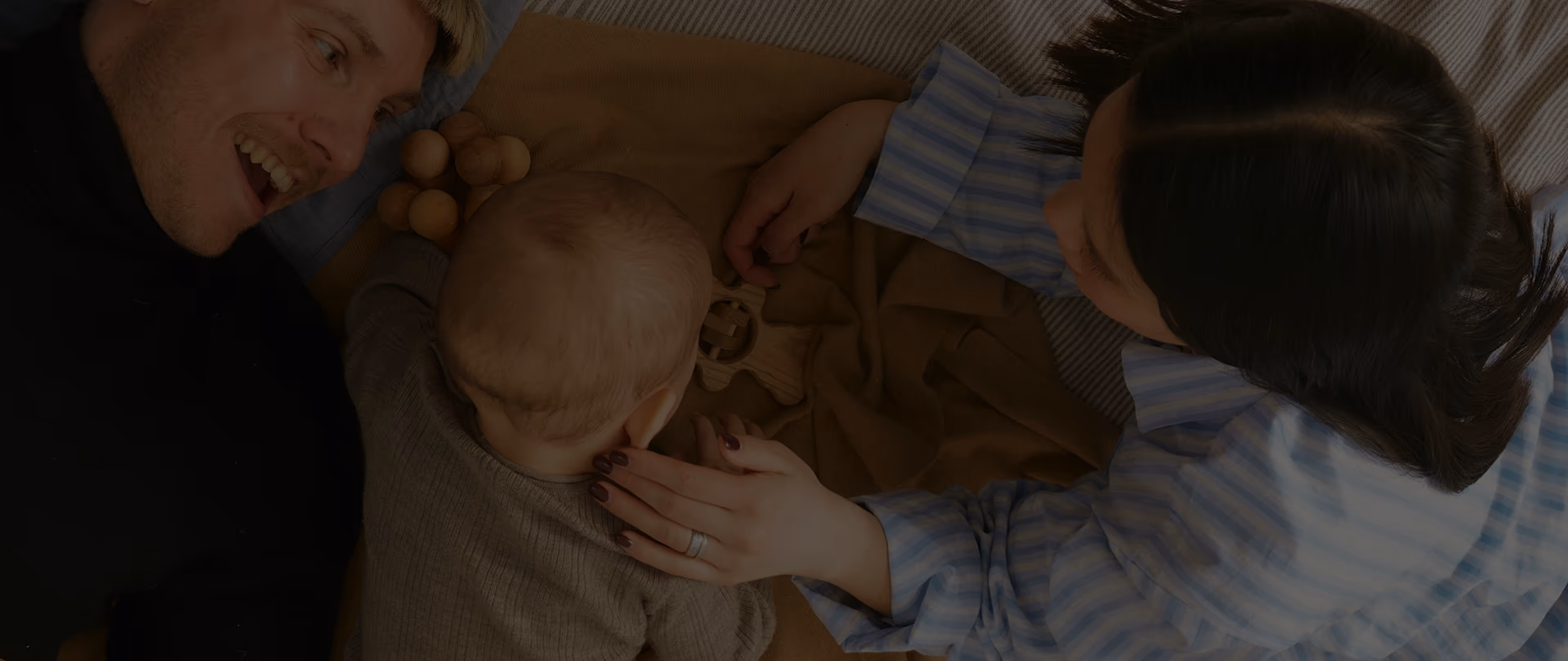 A baby lies between two adults on a blanket, playing with wooden toys.