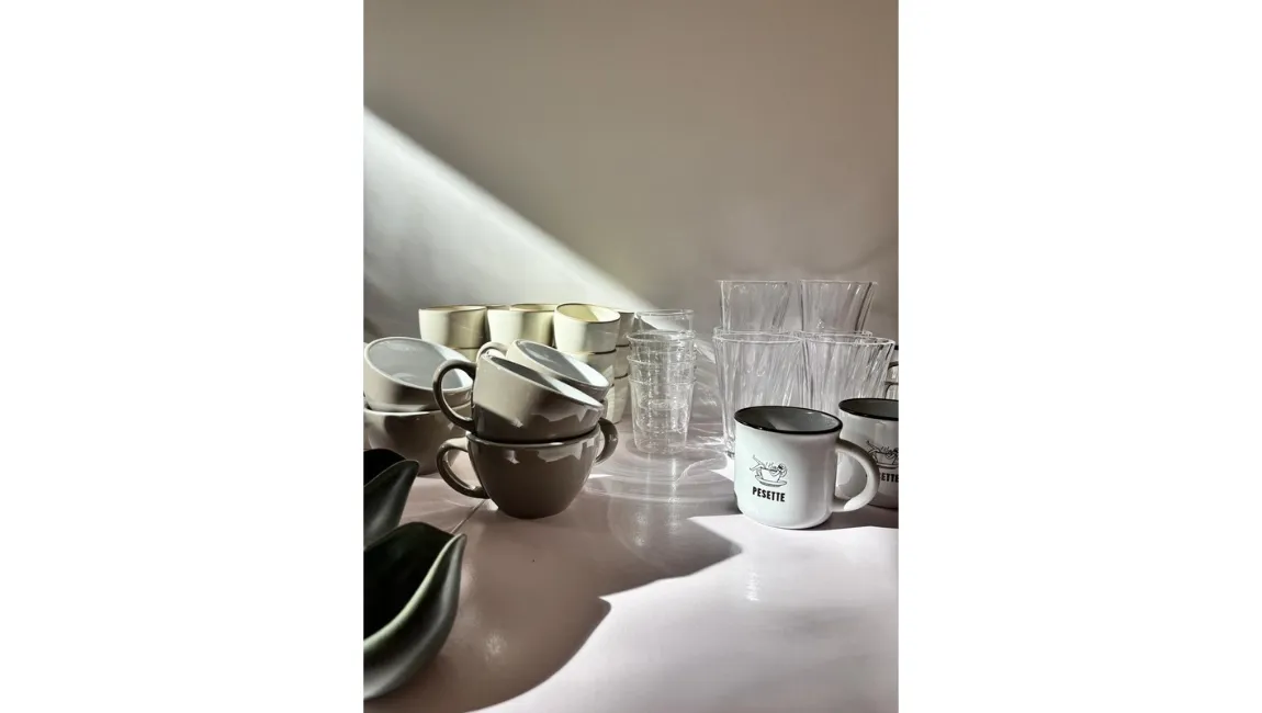 Des tasses en céramique blanches et grises empilées à côté de verres transparents et de mugs blancs avec le logo 'Pesette' sur une surface blanche éclairée par la lumière naturelle.
