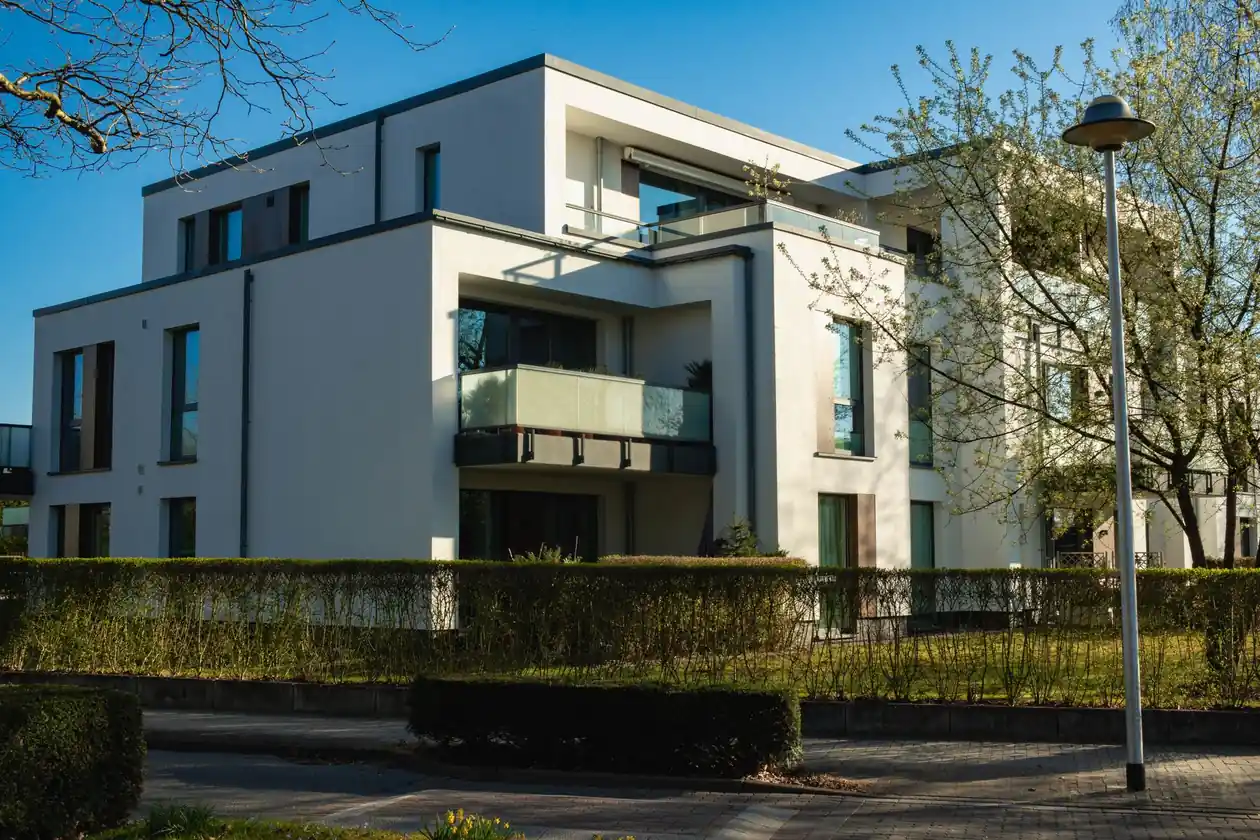 Modern white three-story apartment building with large windows, glass balconies, surrounded by trimmed hedges and trees under a clear blue sky.