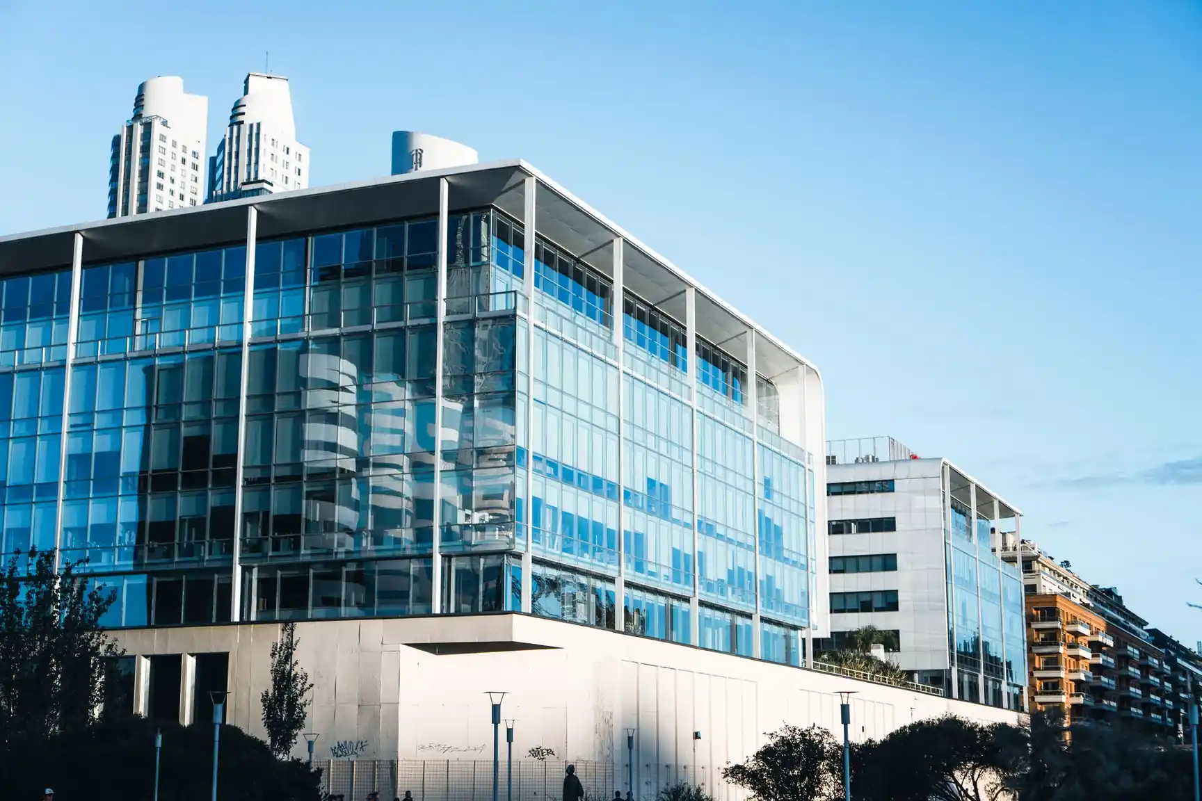 Modern glass and concrete office buildings with reflections of nearby skyscrapers against a clear blue sky.