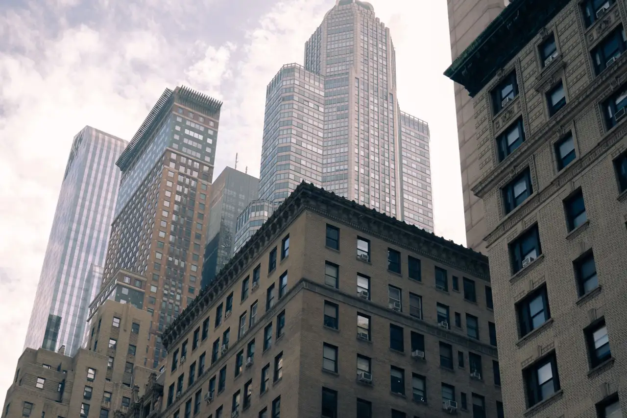 View looking upward at several tall urban buildings with varying architectural styles, including modern glass skyscrapers and older brick structures, under a partly cloudy sky.