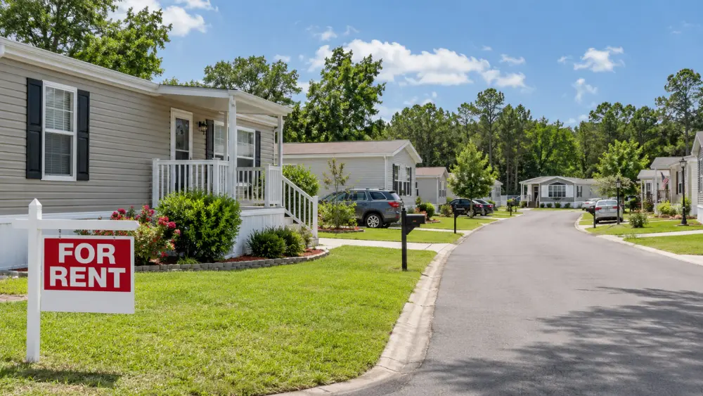 A residential street lined with manufactured homes and green lawns under a blue sky with clouds; a prominent red and white 'For Rent' sign stands on the grass near the sidewalk in front of the nearest home.