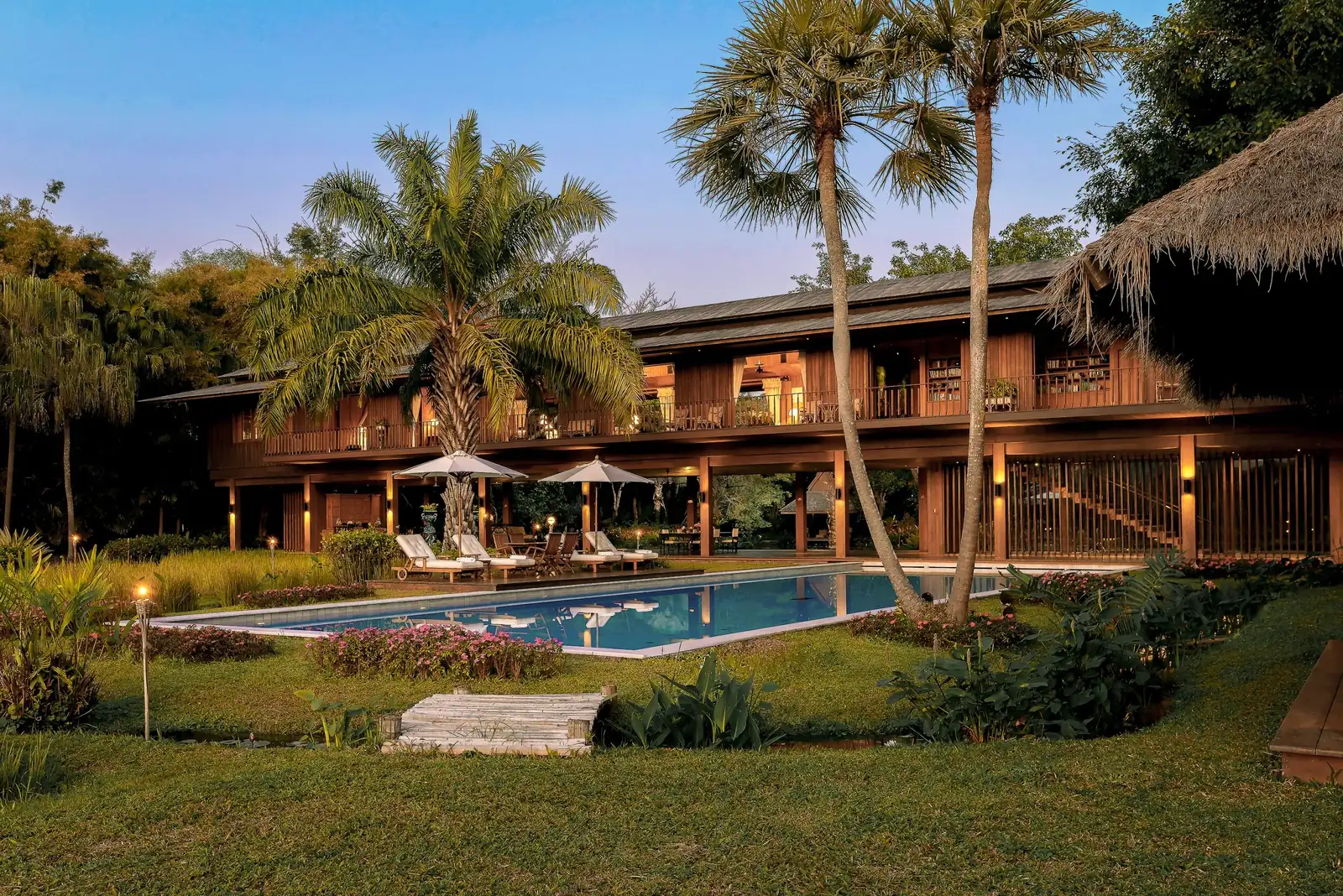 The Tatvani estate at dusk: a two-storey timber house raised above a long pool, framed by palms.