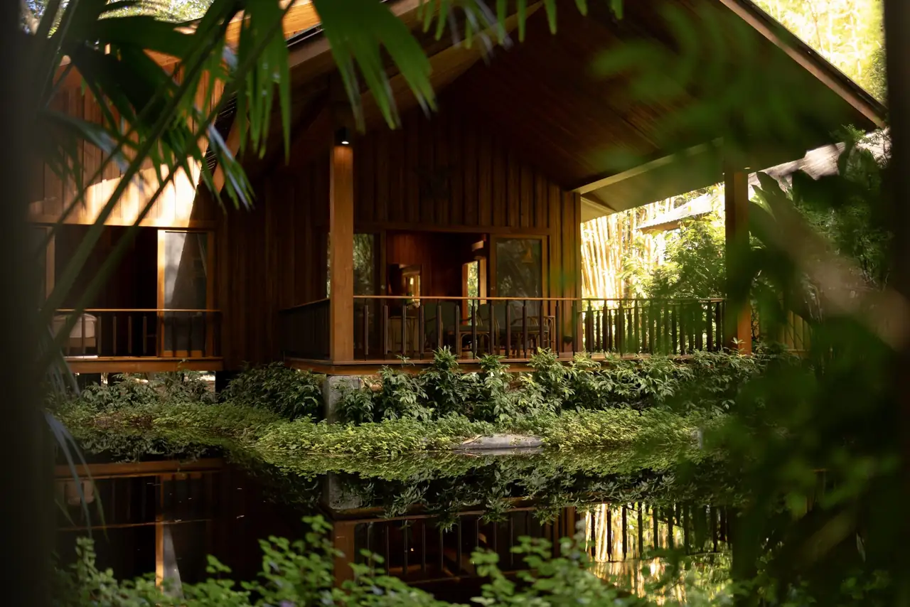 A timber villa reflected in a still pond, seen through leaves.