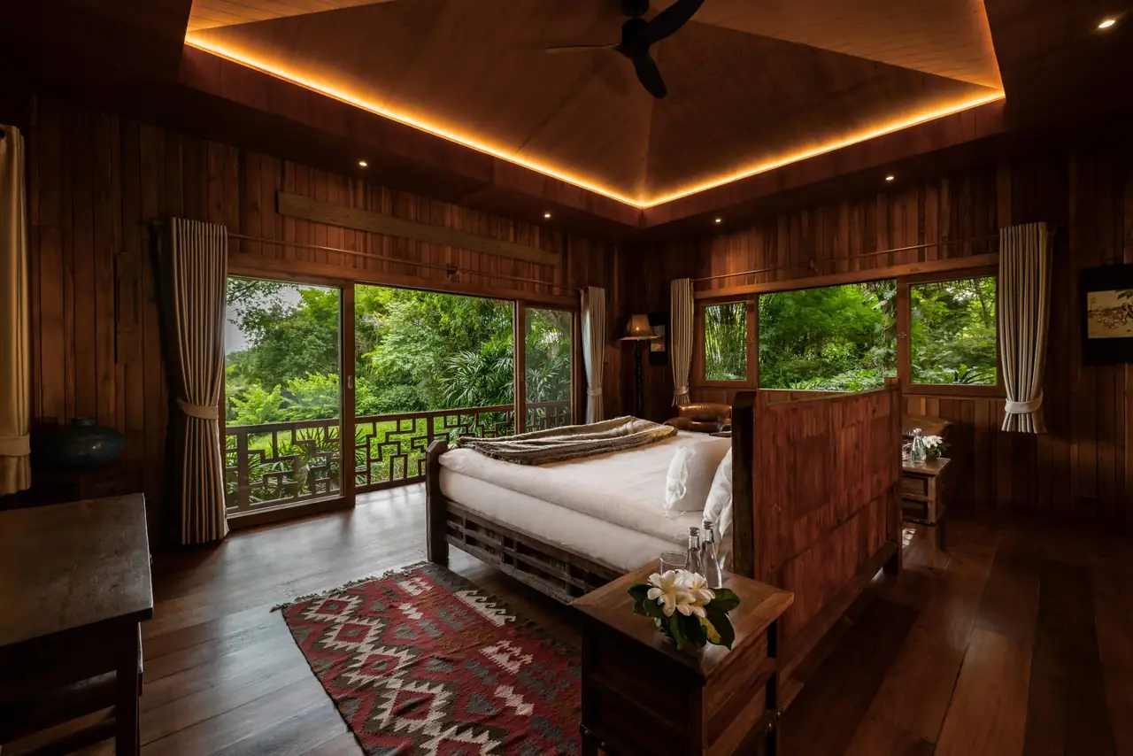 A suite with a lit timber ceiling, a kilim rug and glass doors to a carved balcony over the forest.