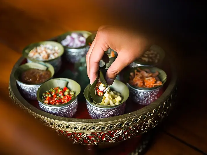 A Thai tray of small bowls holding chopped chilli, ginger, lime and dried shrimp, with a hand assembling a leaf wrap.