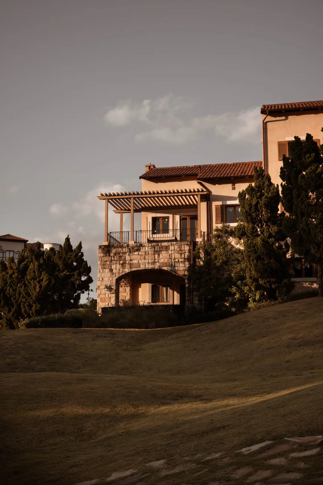 A stone-based villa with a timber pergola, seen across the lawn at golden hour.