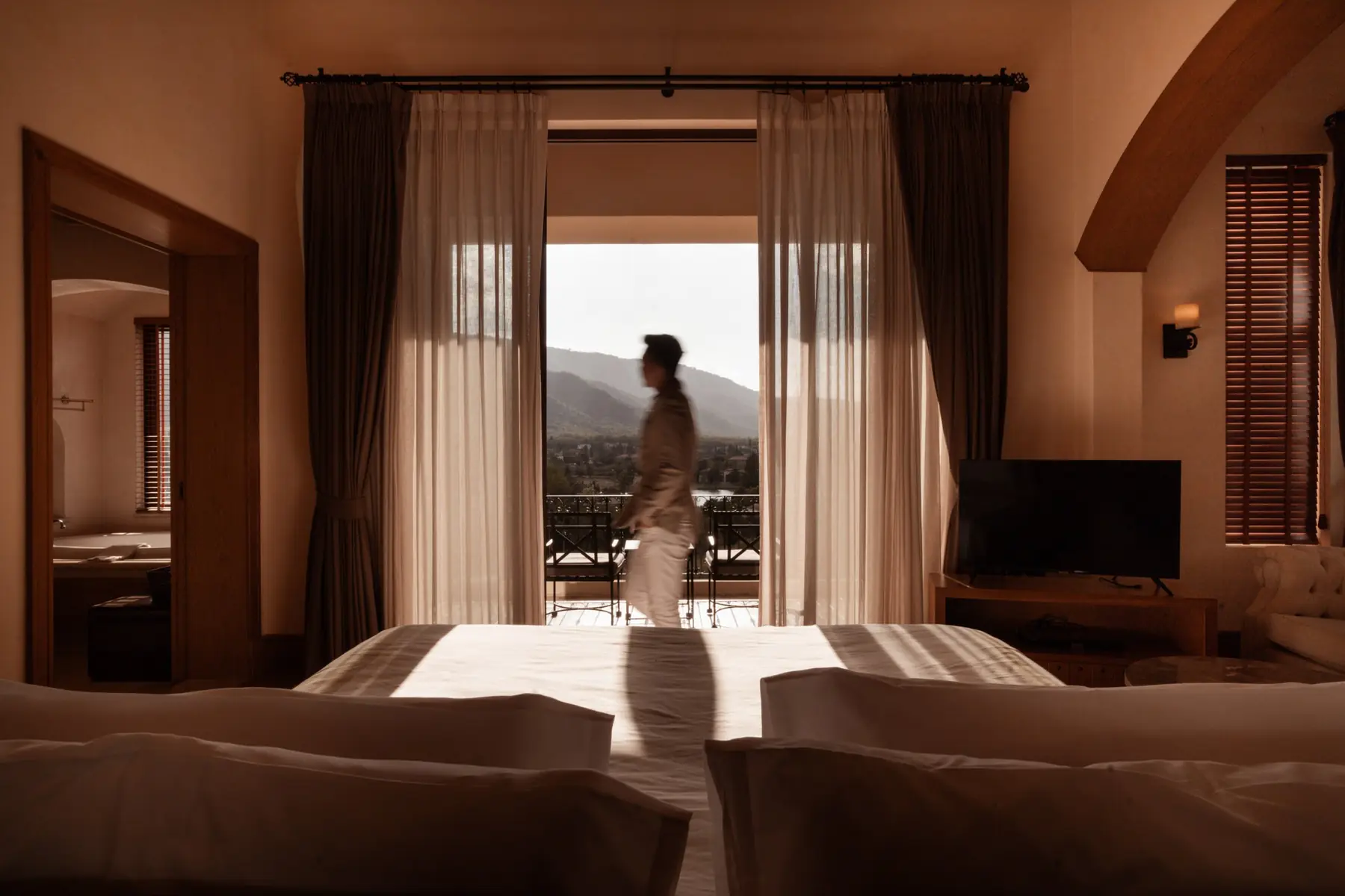 A figure crossing the room in motion, with the balcony and valley framed by the curtains.