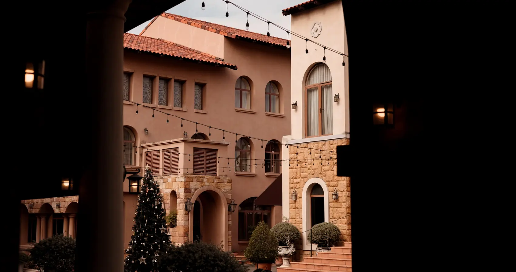 Arched stone and stucco façades with string lights, framed by a dark archway.