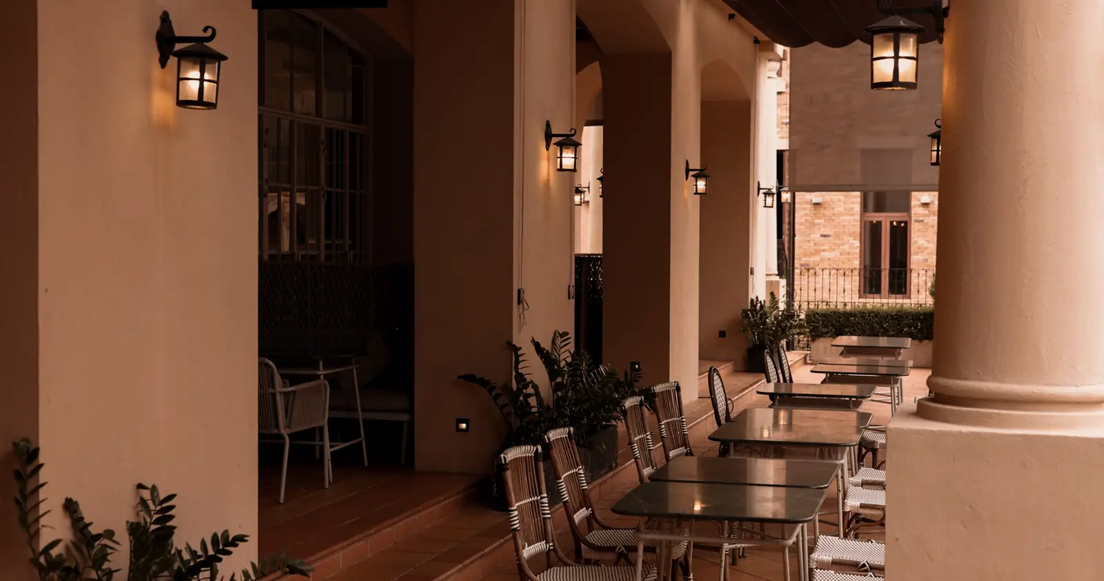 A lantern-lit arcade with café tables and rattan chairs.