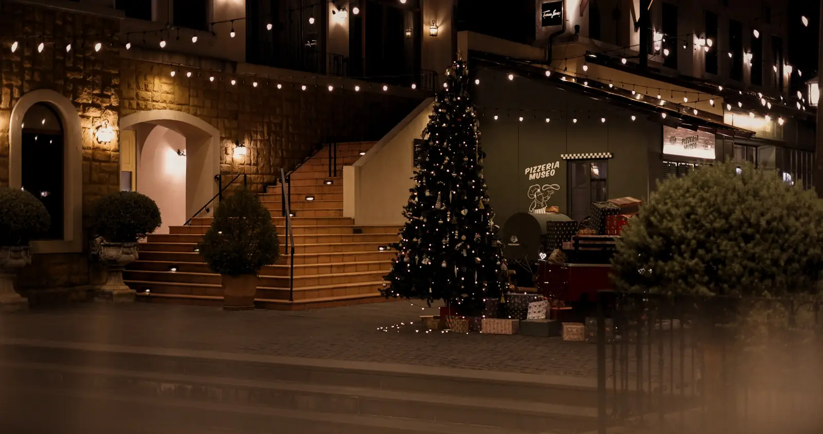 The Christmas tree and gifts on the stairs in Town Square, under string lights at night.