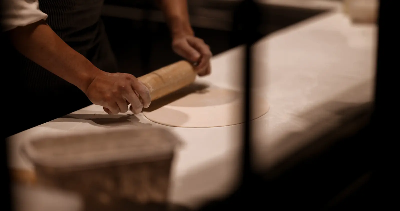 Hands rolling pizza dough with a wooden rolling pin.