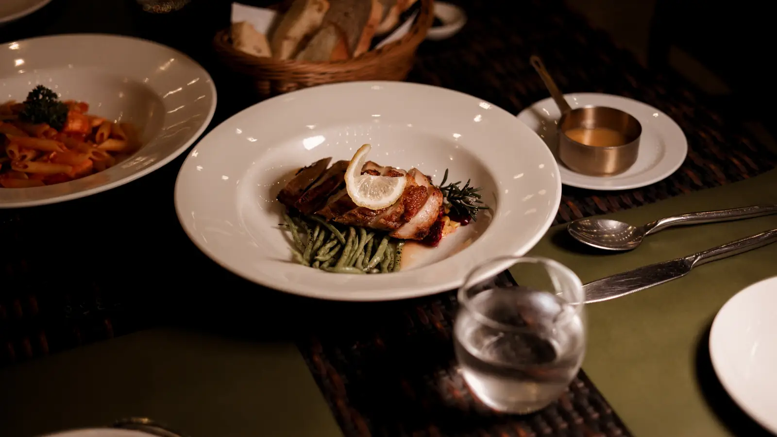 A plated main course with a lemon slice, with pasta behind and a bread basket.