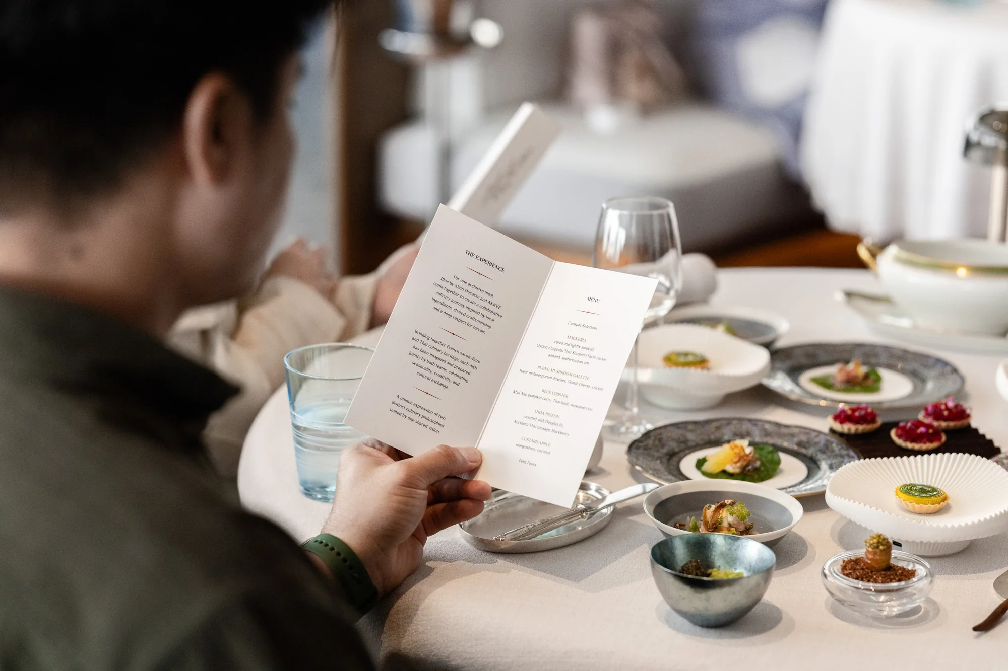 A guest reading the BLUE × AKKEE menu beside a table of plated dishes.