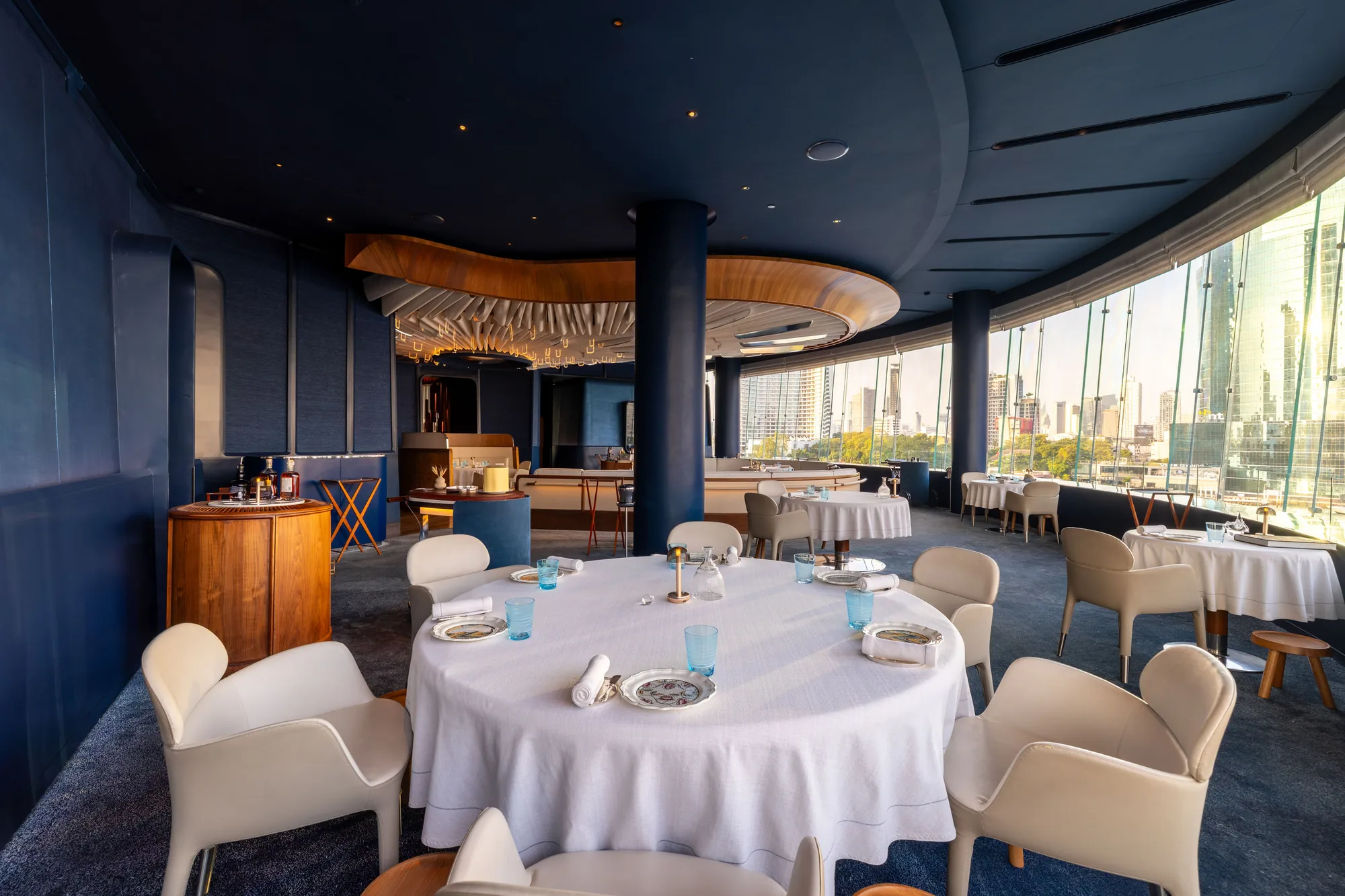 White-linen dining tables framed by deep blue walls, warm wood and panoramic windows.