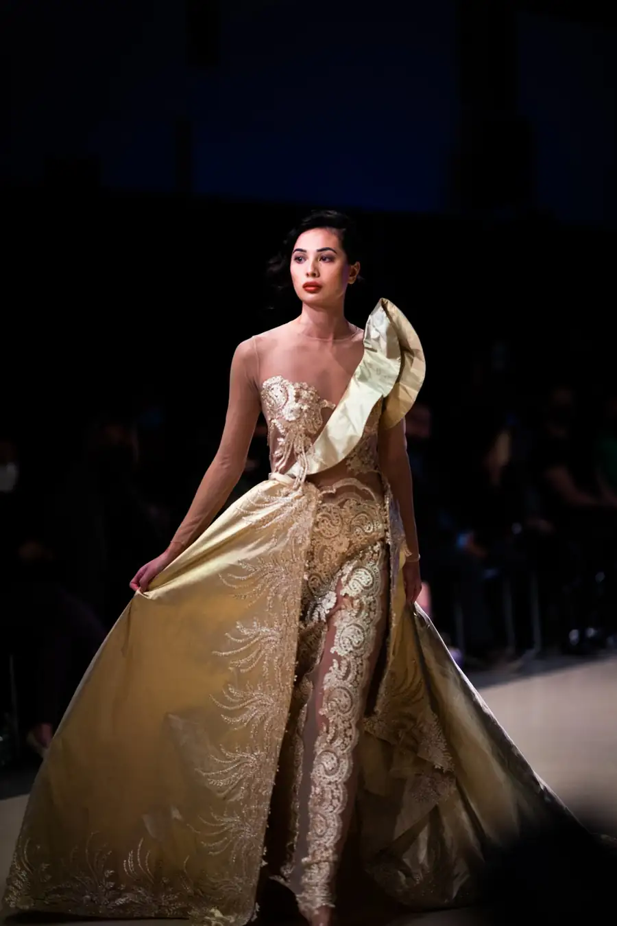 A model on the Vancouver Fashion Week runway in a gold gown with a sculpted shoulder and embroidered lace