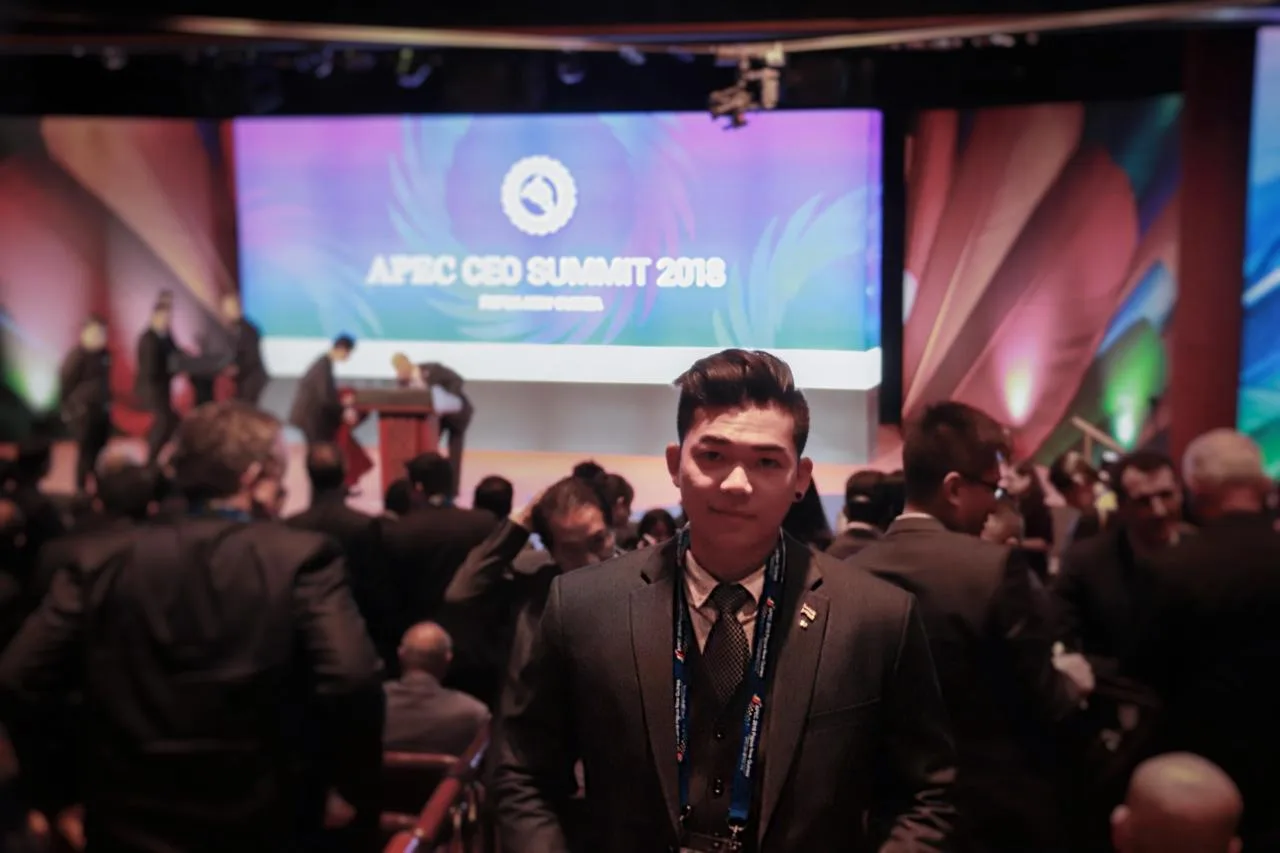 Deejai Riangkrul standing among delegates at the APEC CEO Summit 2018, with the stage behind him.