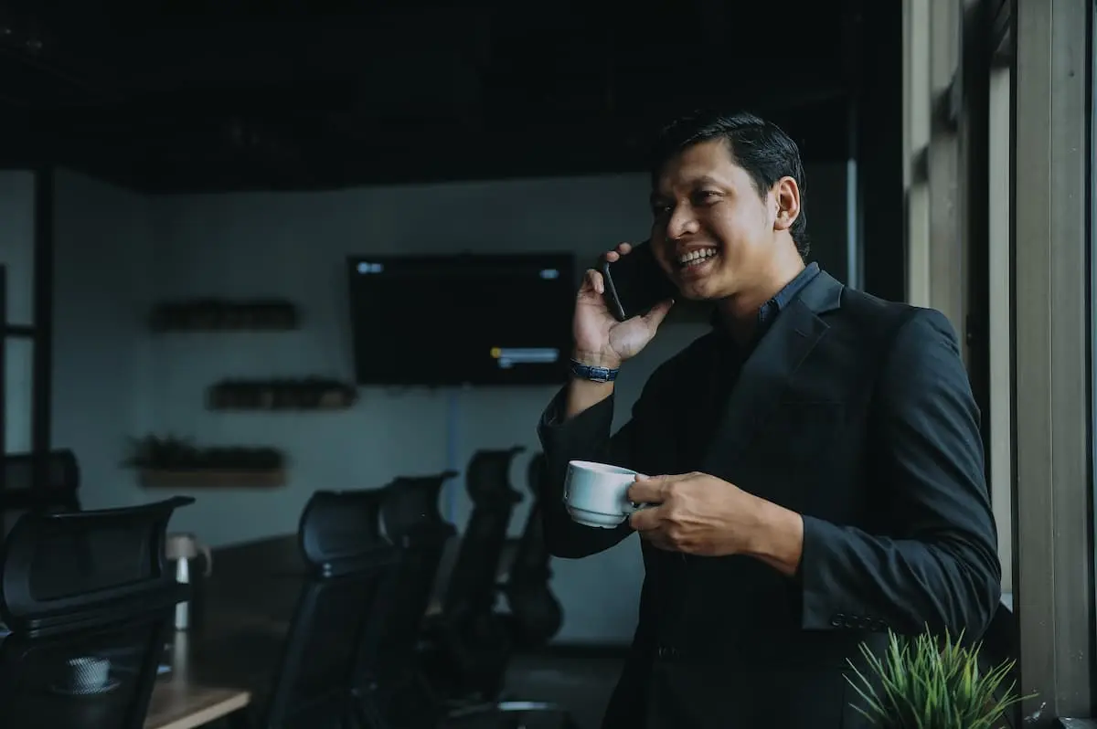 A man is talking on the phone holding a cup of coffee and smiling