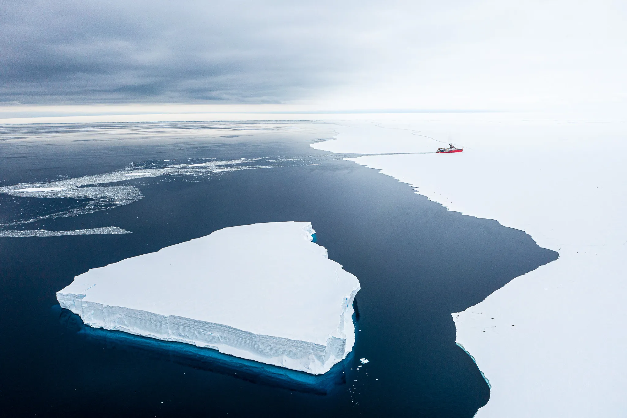 Aerial view of a tabular iceberg in dark open water, with a red research ship at the edge of the sea ice