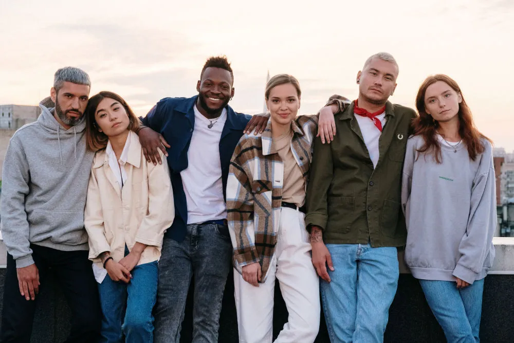 A group of six stylish 30-something men and women standing next to each other on a rooftop, alongside a wall with their arms around each other's shoulders.