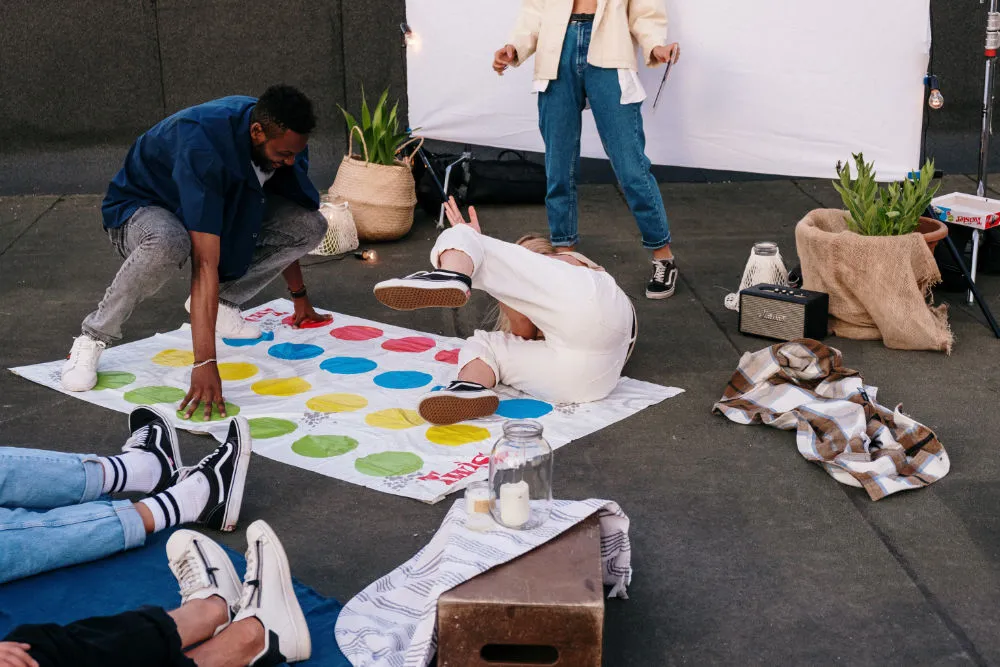 a 30 something woman and man playing the classic game twister on a rooftop while three other people sit and watch.