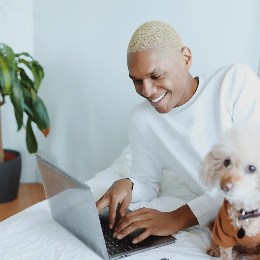 black man in his 30s with blond hair is typing on laptop while laying in his bed in a cozy. there is a small white dog by his side and a plant in the corner of the room.